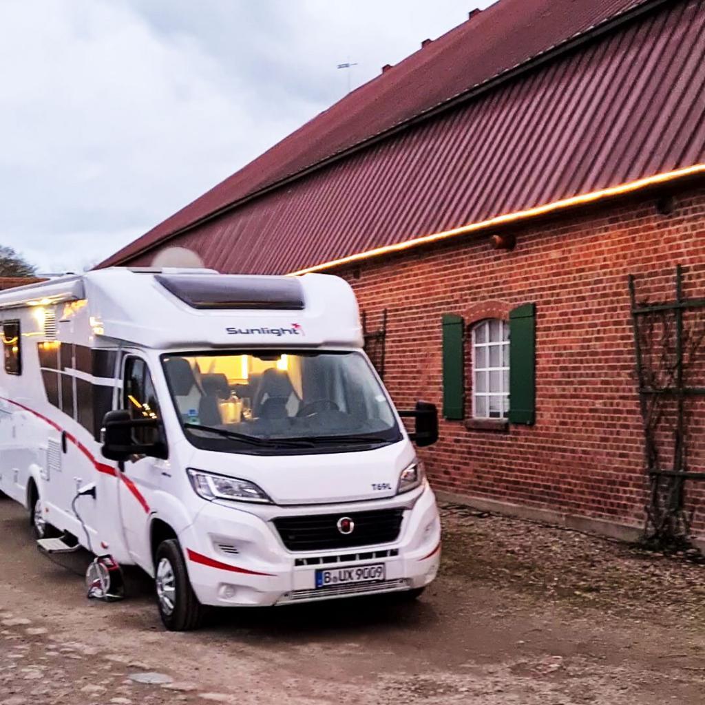 Das Wohnmobil "Rotfuchs" der Camper Vermietung Fuchs und Hase Camper steht auf dem Gutshof vor einer Scheune mit roten Ziegeln und grünen Fensterläden. An der Hauswand sind Pflanzen-ranken zu sehen. Das Wohnmobil ist von innen beleuchtet und strahlt eine gemütliche Atmosphäre beim Wohnmobil Dinner aus.